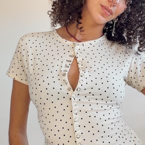 Pode incluir: Uma camisa branca de manga curta com botões e padrão de bolinhas pretas. O texto diz: STUDIO AANYA X DANIELA DOE, BABY TEE BUTTON-UP, PDF SEWING PATTERN + INSTRUCTIONS, SIZE RANGE: XS-XXL, A0, A4 and US Letter.