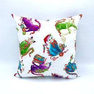 May include: White square pillow with a repeating pattern of whimsical cats in various colors, including purple, green, and blue. The cats are adorned with festive decorations like candy canes, ornaments, and gift boxes, creating a holiday theme.