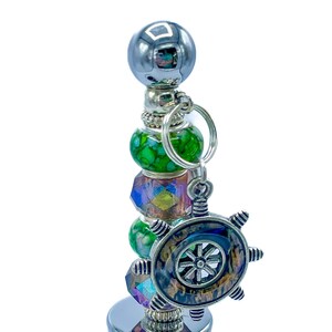 May include: A decorative bottle stopper with a silver finish, featuring a ship's wheel charm. The stopper has green and iridescent beads, and a silver ball at the top. The stopper is designed to fit a wine bottle.