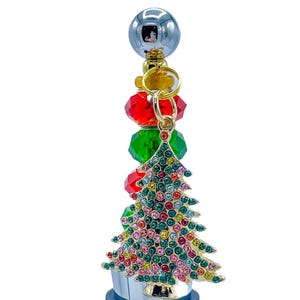 May include: A decorative bottle stopper with a Christmas tree design. The stopper features a gold-colored tree adorned with colorful rhinestones. It is attached to a silver-colored stopper with red and green crystal beads and a silver ball.