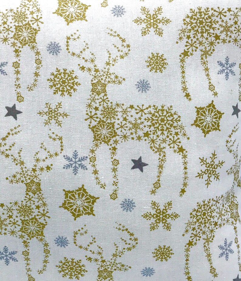 May include: White fabric with a repeating pattern of gold snowflakes and stars. The snowflakes are arranged to form the shape of a reindeer.