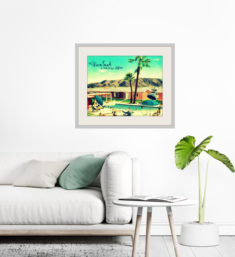 May include: Framed art print featuring a vintage-style illustration of a mid-century modern resort scene. The artwork depicts a swimming pool, palm trees, and a retro building, with the text "the Warm Sands, Palm Springs, California". The print is displayed on a white wall.