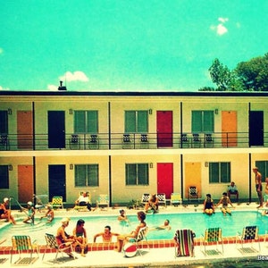 Pode incluir: Um motel vintage com uma piscina na frente. O motel tem um exterior branco com portas e janelas coloridas. As pessoas est&atilde;o nadando e relaxando perto da piscina.