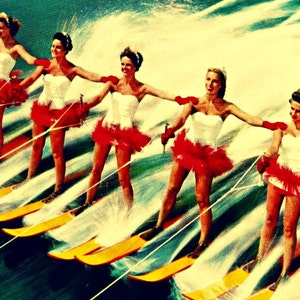 May include: Five women in white dresses with red ruffles are water skiing in a line. They are all smiling and holding onto ropes attached to a boat. The water is blue and there is a lot of spray from the skis.