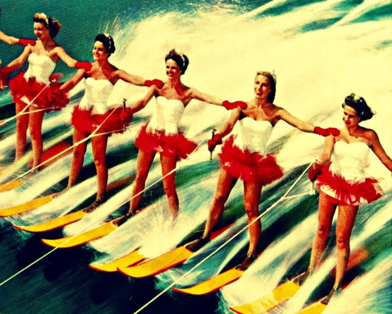 May include: Five women in white dresses with red ruffles are water skiing in a line. They are all smiling and holding onto ropes attached to a boat. The water is blue and there is a lot of spray from the skis.