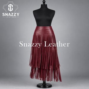 May include: A deep burgundy leather skirt featuring a high waistband and multiple layers of long, thin fringe that hang down to create a textured, flowing appearance. The skirt is displayed on a black mannequin with the text Snazzy Leather.