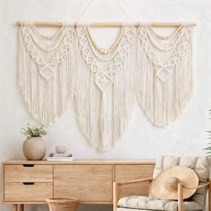 May include: A cream-colored macrame wall hanging with a wooden dowel and beaded accents. The design features geometric patterns and long fringe, creating a bohemian aesthetic. The wall decor is displayed above a wooden cabinet.