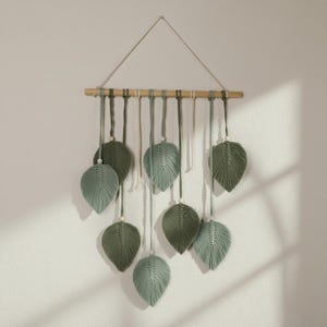 Könnte beinhalten: Makramee-Wandbehang mit blattförmigen Designs in Grün- und Blaugrüntönen. Die Blätter sind an einem natürlichen Holzdübel mit einer Schnur zum Aufhängen befestigt. Ein minimalistischer, von der Natur inspirierter Einrichtungsgegenstand.