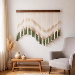 May include: A decorative wall hanging with a wooden bar, cream-colored strings, wooden beads, and green and cream tassels. The wall decor is displayed on a white wall, with a wooden bench, vases, and a gray armchair in the foreground.