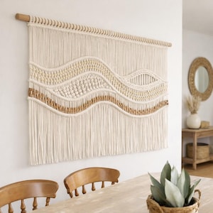 May include: A cream-coloured macrame wall hanging with a wooden dowel. The design features woven patterns and a wavy, textured design with tan and light brown accents. The piece hangs on a white wall, adding a bohemian touch to the room.