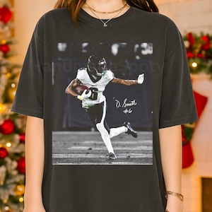 May include: Dark gray t-shirt featuring a black and white image of a football player in action, holding a football. The player is wearing a helmet and uniform. The shirt has the player's name and number on it.