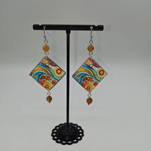 Peut inclure: Une paire de boucles d'oreilles pendantes avec des pendentifs en forme de losange arborant un motif abstrait coloré de tourbillons et de fleurs bleus, jaunes et orange. Chaque boucle est ornée d'une petite perle ronde ambrée au-dessus et en dessous.