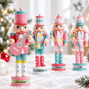 May include: Four wooden nutcracker figurines painted in pastel pink, blue, and white. Each stands on a round base with a name printed on it: Eleanor, Agnes, Owen, and Lila. They feature white hair, beards, and various accessories like a guitar.
