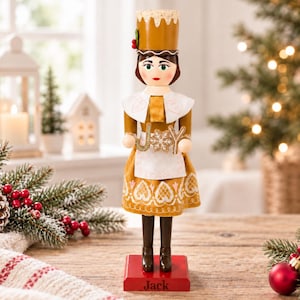 May include: A nutcracker figure wearing a golden dress with white apron and hat, holding a sign that reads JOY. The figure has brown hair, green eyes, and stands on a red rectangular base with the name Jack printed in black on the front.