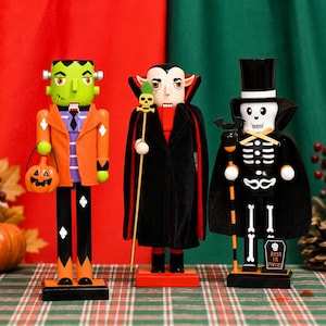 May include: Three wooden nutcrackers dressed as Halloween characters: a green Frankenstein monster holding a pumpkin bucket, a vampire in a black cape holding a skull staff, and a skeleton in a top hat and cape holding a bat staff with a tombstone.