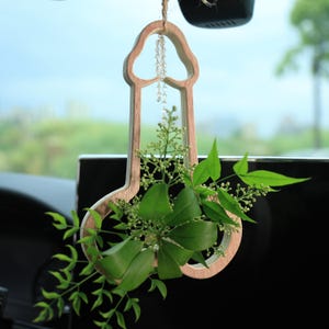 Könnte beinhalten: Ein hölzerner Autoanhänger in Form eines stilisierten Phallus, gefüllt mit grünen Blättern und kleinen weißen Blüten. Der Anhänger hängt an einer Schnur aus Naturfaser und hat eine Kette aus klaren Perlen. Der Hintergrund ist unscharf und zeigt einen Autoinnenraum und die Außenansicht.