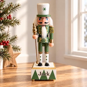 May include: A wooden nutcracker figure wearing a green uniform with gold accents, a white hat featuring a green snowflake, and white hair and beard. The nutcracker holds a gold staff and stands on a white base with green triangles and the name Jack.