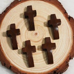 May include: Five small, dark brown wooden crosses are arranged on a light brown, circular wooden surface. The crosses have a simple, classic design. The wood grain of the surface is visible, adding texture to the image.