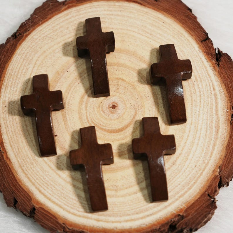 May include: Five small, dark brown wooden crosses are arranged on a light brown, circular wooden surface. The crosses have a simple, classic design. The wood grain of the surface is visible, adding texture to the image.