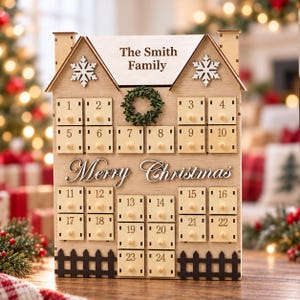 May include: A light-coloured wooden house-shaped advent calendar featuring twenty-four small drawers with knobs, numbered one through twenty-four. The front displays the text "The Smith Family" and "Merry Christmas," decorated with two white snowflakes and a small green wreath.