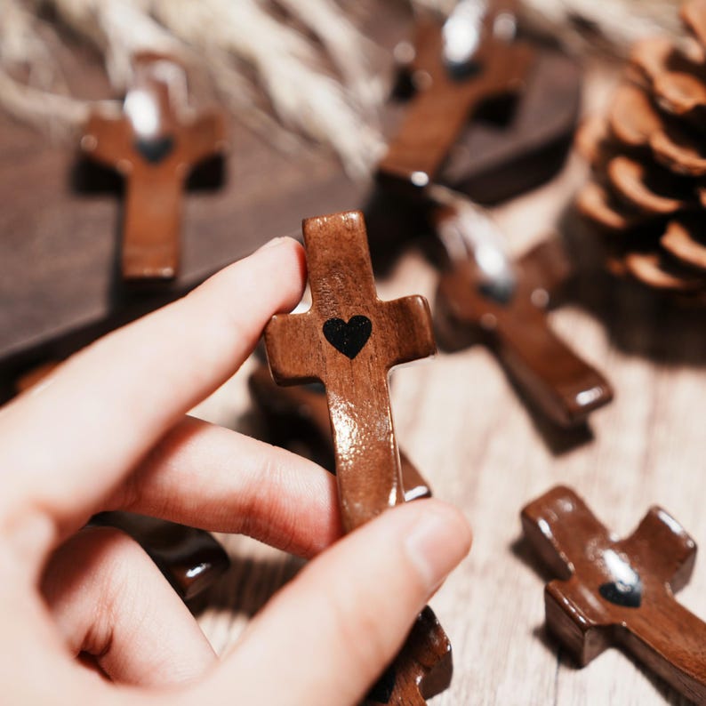 May include: Wooden crosses with a dark brown finish and a black heart in the center. The crosses are held in a hand, with several others scattered on a wooden surface. The crosses are a religious item.
