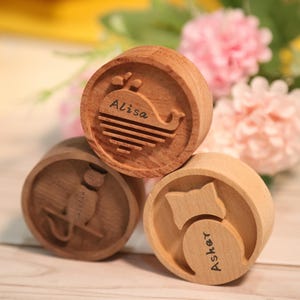 Op de afbeelding: Drie houten stempels met gegraveerde ontwerpen. Eén stempel toont een walvis met de naam "Alisa", een andere een kat met "Caitlin" en de laatste een kat met "Asher". De stempels zijn lichtbruin en gestapeld.