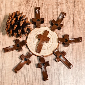Könnte beinhalten: Eine Ansammlung brauner Holz-Kreuze, jedes mit einem kleinen schwarzen Herzdetail, auf einer Holzoberfläche angeordnet. Daneben liegt ein Tannenzapfen. Die Kreuze variieren leicht in Form und Größe und wirken rustikal und handgefertigt.