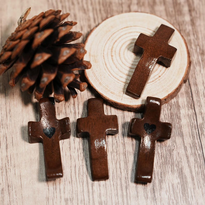 May include: A collection of small, wooden crosses in varying positions. The crosses are a rich, dark brown color, with a glossy finish. Some crosses have a small, black heart detail. A pine cone and a wooden slice are also present.