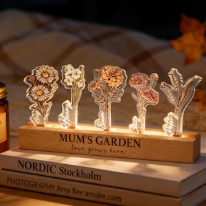Op de afbeelding: Een houten basis met de tekst "MUM'S GARDEN love grows here" ondersteunt vijf heldere acryl bloemuitsparingen, elk met een ander bloemontwerp en een naam. De basis staat op een stapel boeken. De bloemen worden verlicht door een warm licht.