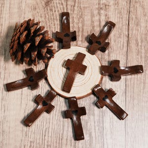 May include: A collection of eight brown wooden crosses, each with a small black heart detail. The crosses are arranged around a wooden slice, with a pine cone on the side. The background is a light brown wooden surface.