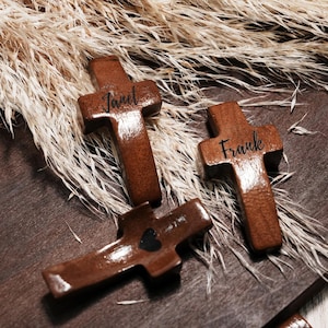 May include: Three wooden crosses with a dark brown finish. Two crosses have names engraved: "Janet" and "Frank". The third cross has a black heart. The crosses are arranged on a wooden surface with dried plants in the background.