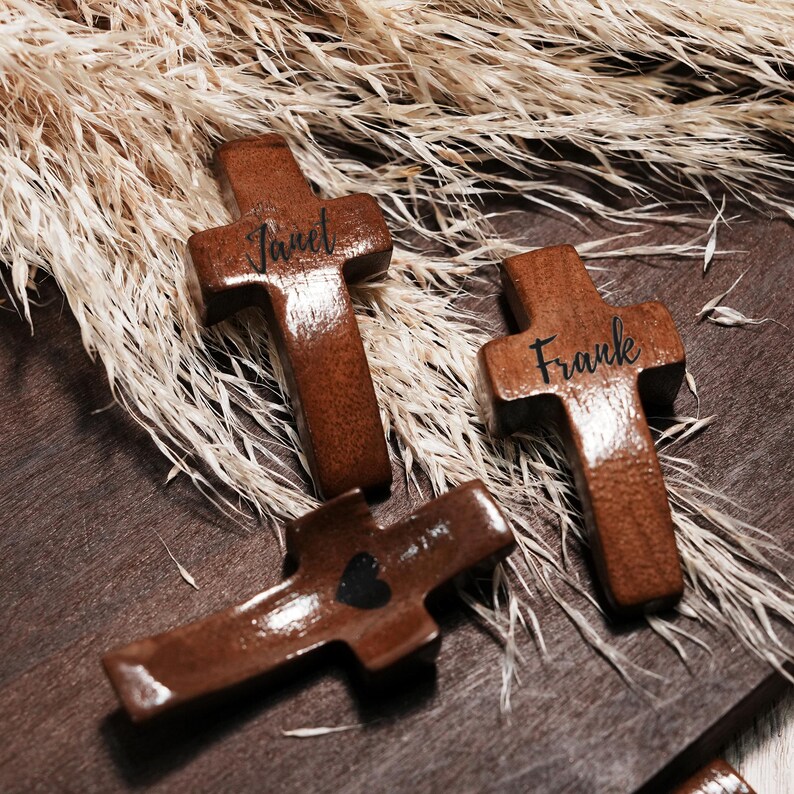May include: Three wooden crosses with a dark brown finish. Two crosses have names engraved: "Janet" and "Frank". The third cross has a black heart. The crosses are arranged on a wooden surface with dried plants in the background.