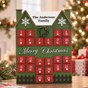 Könnte beinhalten: Ein grüner, hausförmiger Adventskalender aus Holz mit vierundzwanzig roten Schubladen, die jeweils mit einer weißen Zahl versehen sind. Der Text "The Anderson Family" und "Merry Christmas" ist aufgedruckt, ergänzt durch weiße Schneeflocken.