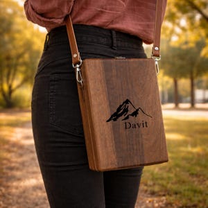 May include: A rectangular wooden crossbody bag with a smooth, dark brown grain finish. The front features a black graphic of a mountain range above the text "Davit". It includes a thin, adjustable brown leather strap attached with silver metal hardware.