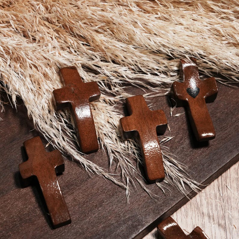May include: Several small, wooden crosses with a glossy finish are arranged on a dark wooden surface. The crosses are a rich brown color and have a simple, classic design. Dried, beige-colored plants are in the background.