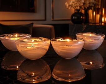 May include: Four Kosta Boda glass bowls arranged in a group, each featuring a swirling white and clear glass pattern. Each bowl contains a lit tea light candle, casting a warm glow. The brand name Kosta Boda is etched on the front of each bowl.