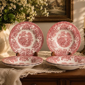 Può includere: Un set di quattro piatti in ceramica con un motivo a trasferimento rosso e bianco. Il design include una scena paesaggistica centrale circondata da intricati bordi floreali e bordi smerlati. Due piatti sono in piedi, due sono impilati.