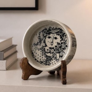 Peut inclure: Un bol en céramique décoratif avec une illustration en noir et blanc d'un visage de femme et de motifs floraux. Le bol est présenté sur un support en bois. Le bol a un fond blanc et un bord noir.