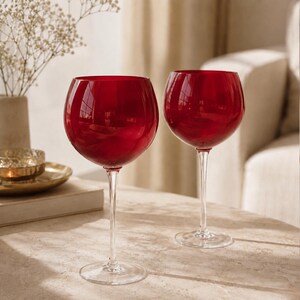 May include: Two wine glasses featuring deep red, rounded bowls and clear, slender stems. The glasses are crafted from smooth, translucent glass, showcasing a vibrant crimson hue that catches the light, supported by stable, circular clear glass bases.