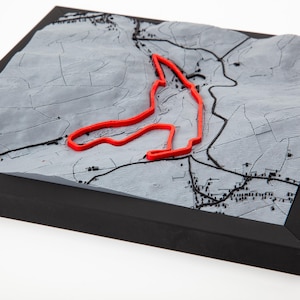 Puede incluir: Un mapa 3D enmarcado con un contorno rojo de una pista de carreras sobre un fondo topográfico gris. El mapa está enmarcado en un marco negro, mostrando el diseño y el paisaje de la pista. Una pieza única para los aficionados al automovilismo, en cm.