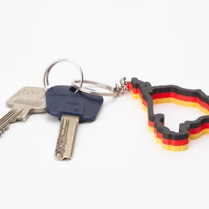 Op de afbeelding: Een sleutelhanger met een Duits vlagontwerp, met zwarte, rode en gele strepen, bevestigd aan een sleutelring met twee sleutels. De sleutelhanger heeft de vorm van een kaartomtrek.