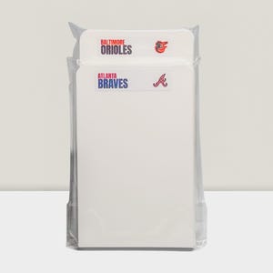 May include: A package of white notepads featuring the logos of the Baltimore Orioles and Atlanta Braves baseball teams. The team names and logos are in red, blue, and white. The notepads are sealed in clear plastic.