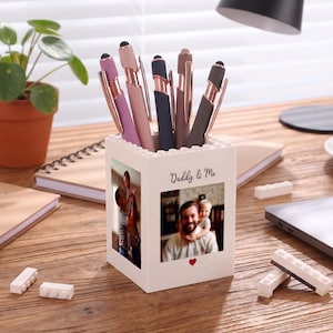 May include: A white square pen holder constructed from building blocks, featuring two photo inserts on the front and side. The front panel displays the text "Daddy & Me" above a photograph of an adult and a child, with a small red heart below.