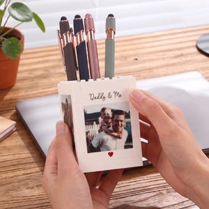 May include: A white square pen holder constructed from interlocking building blocks, featuring a printed photograph of a man and a child with the text Daddy & Me and a small red heart. Four stylus pens in brown, navy, rose gold, and gray are inside.