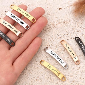 May include: A collection of small, rectangular metal tags in gold, silver, rose gold, and black finishes. Each tag features rounded ends with holes and is engraved with various phrases including You are amazing, BRING IT ON!, Gib nicht auf, I believe in you, MAKE MY DAY, Du bist stark, I trust in you, and Follow your heart.