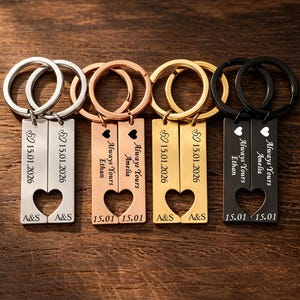May include: Four pairs of rectangular metal keychains in silver, rose gold, gold, and black tones. Each pair features a cutout heart shape, engraved text including names like Ethan and Amelia, dates like 15.01.2026, and the initials A&S.