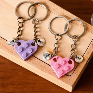 May include: Two keychains with heart-shaped charms made of purple and pink building blocks. Each keychain has a silver ring, chain, and a small, round charm with a letter. The keychains are on a wooden surface.