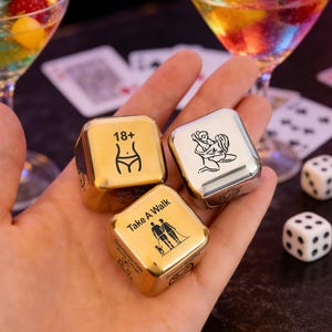 May include: Three metallic dice with various illustrations and text, including "18+", a couple, and a couple embracing. The dice are held in a hand, with cocktails and playing cards in the background.