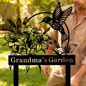 Op de afbeelding: Zwart metalen tuinbord met een kolibrie en bloemenontwerp. Het bord leest "Grandma's Garden" in gouden letters. De kolibrie is in vlucht en de bloemen zijn lelies. Het bord is ontworpen om in een tuin te worden geplaatst.
