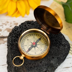 May include: A brass compass with an open lid, showing the compass rose and directional markings. The compass sits on a dark, textured surface. The lid is engraved with the word "HAYDEN" and a mountain range.